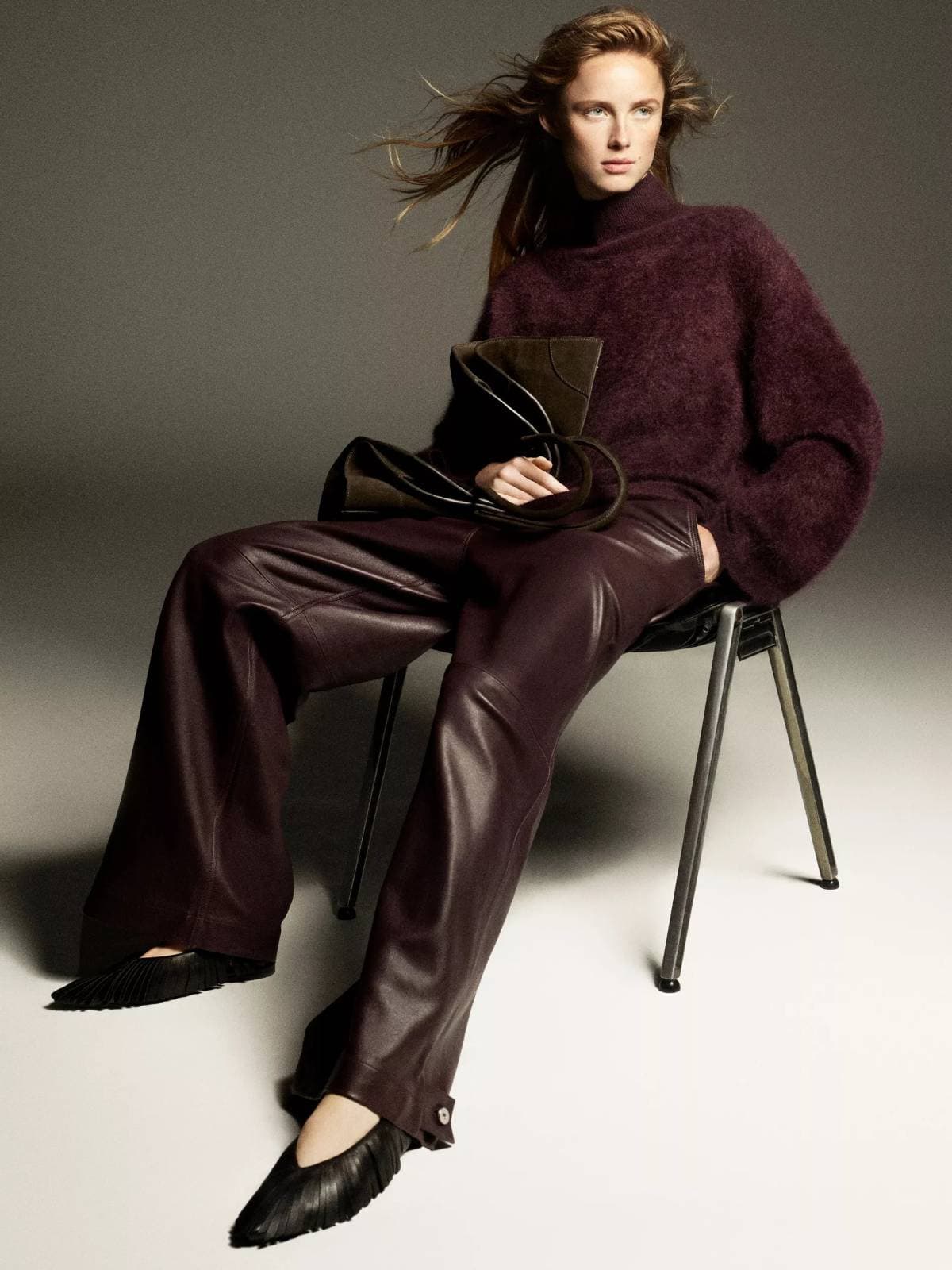 Model seated on a chair wearing a textured burgundy sweater and leather trousers while holding a dark leather handbag.