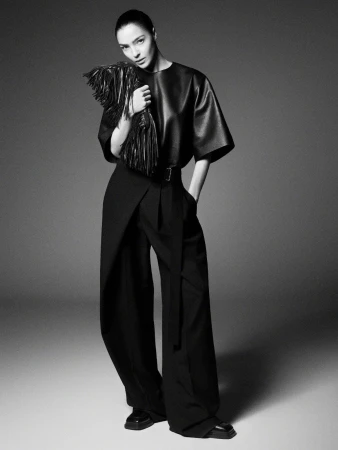 Model wearing dark leather top and wide trousers holding fringed bag against grey gradient studio background