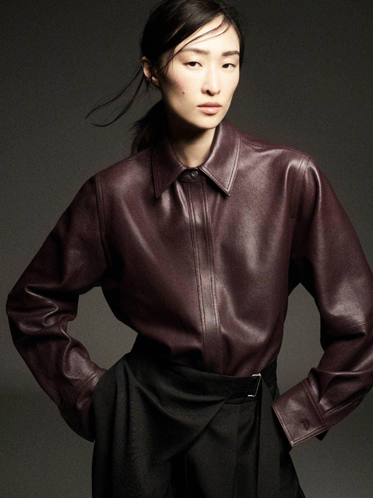 Model wearing a deep burgundy leather shirt and dark skirt posing against a neutral grey studio backdrop