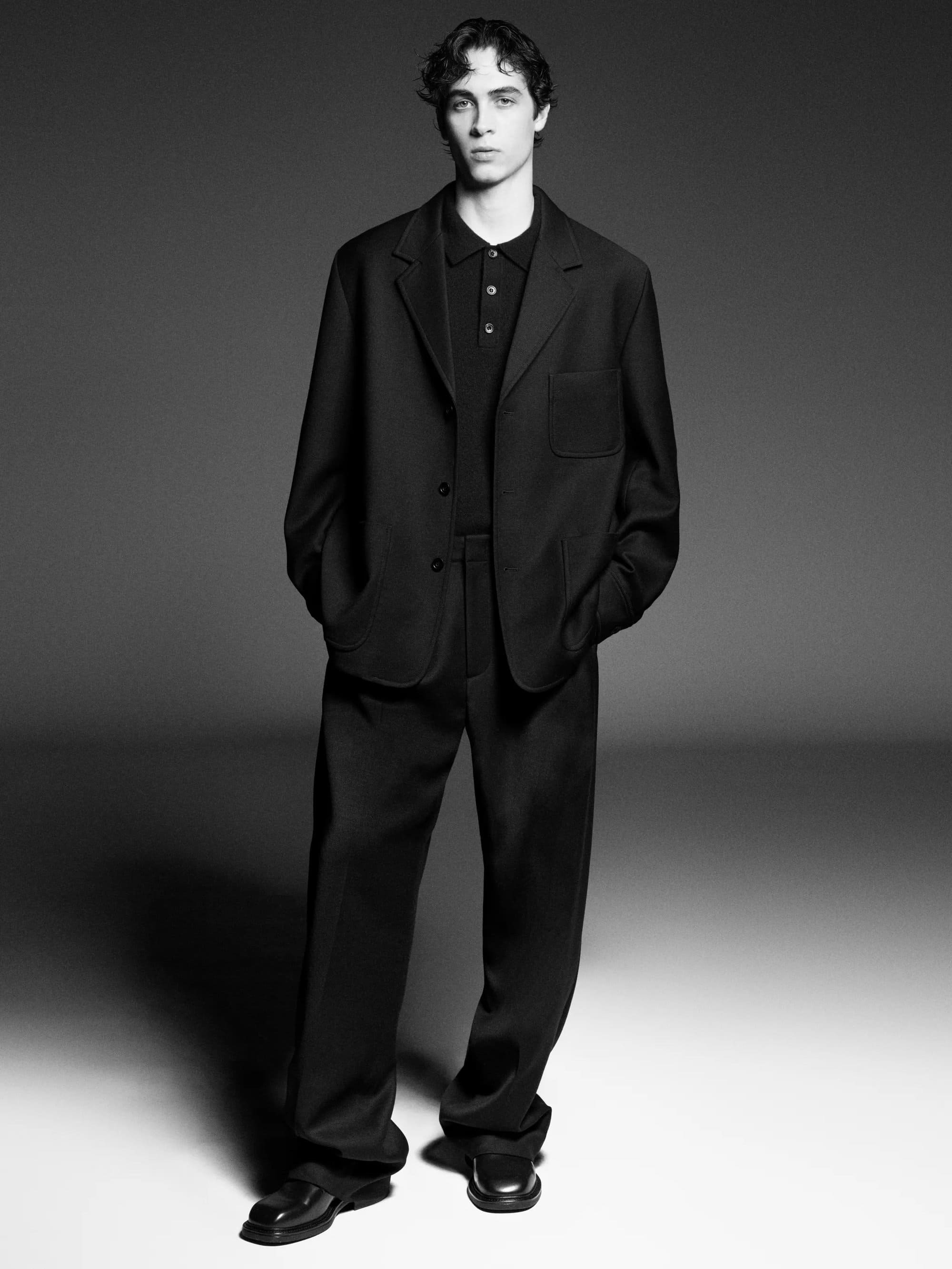 Man wearing a black blazer, polo shirt, and trousers standing against a gradient studio background.
