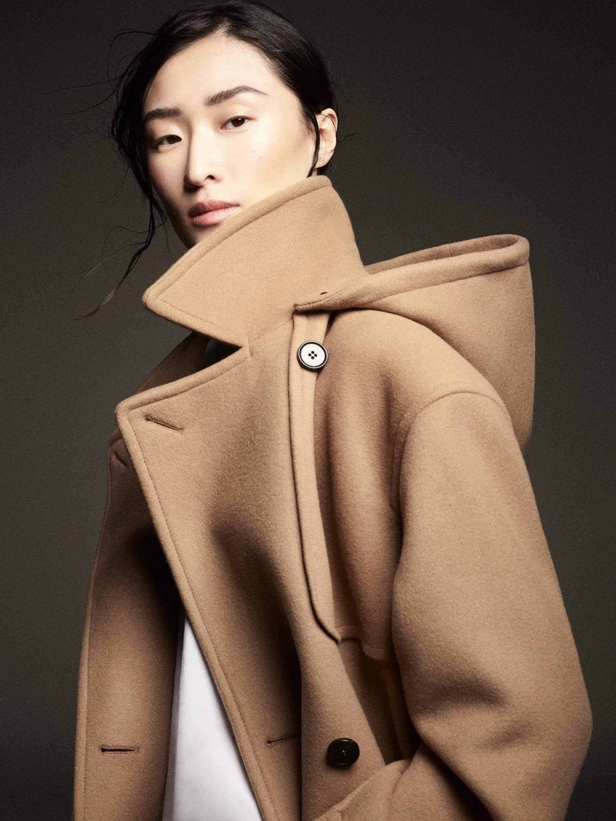 Model wearing a camel hooded coat with dark hair blowing softly against a dark background for COS Fall 2024 campaign.