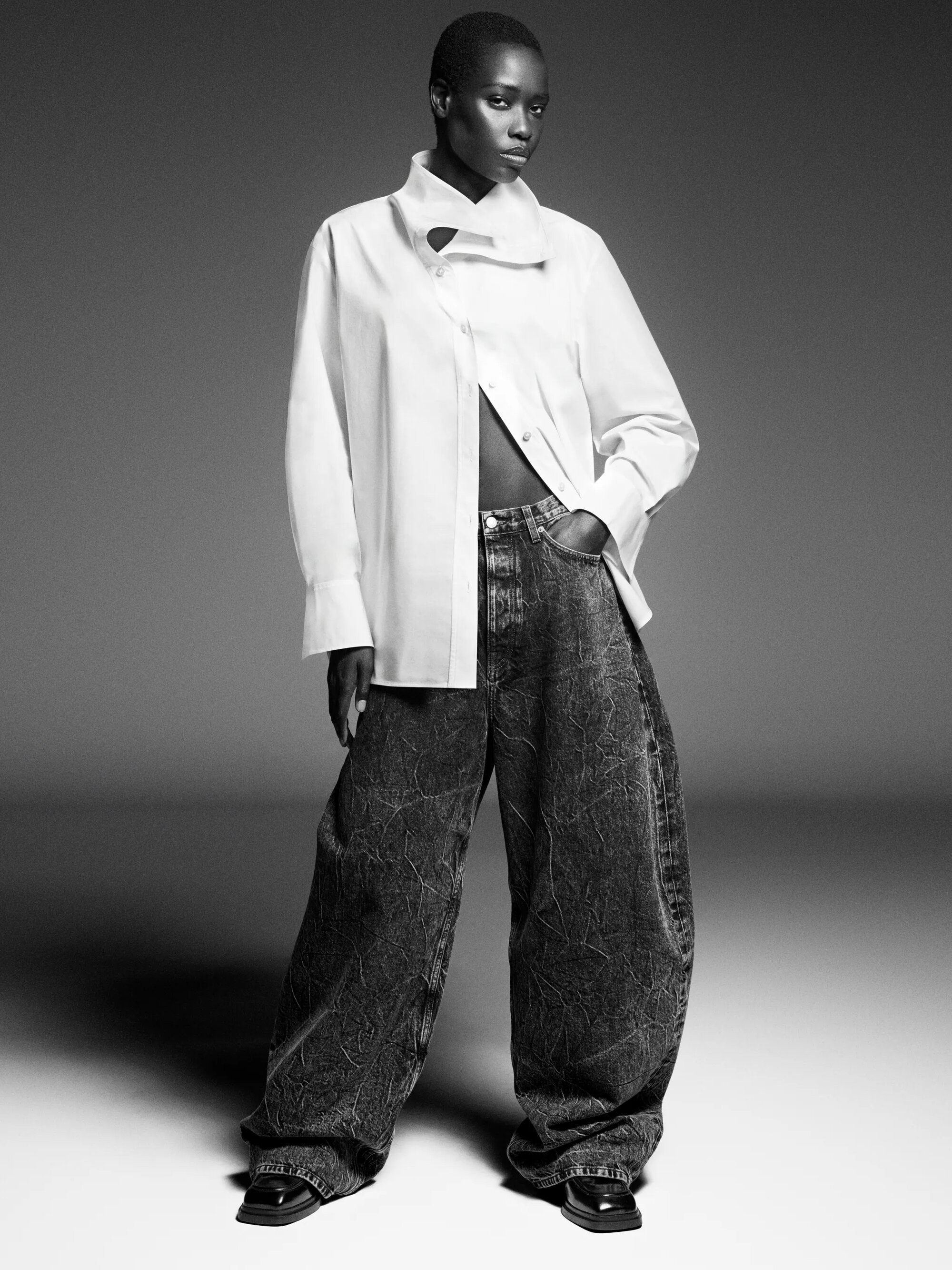 Model in white buttoned shirt and wide dark jeans standing against a plain grey backdrop for COS.