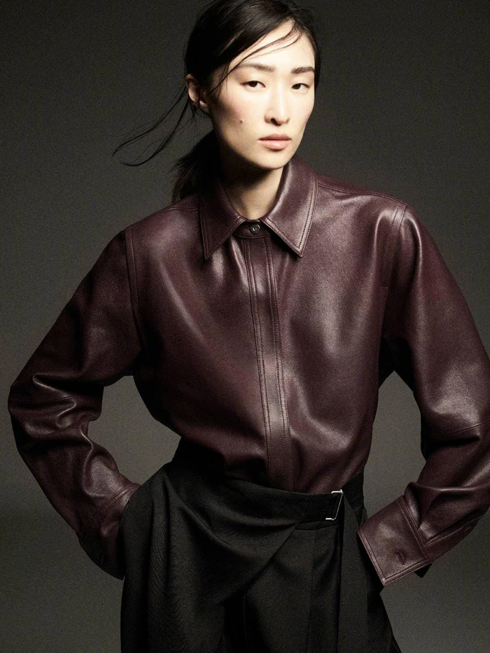 Model wearing a deep burgundy leather shirt and dark skirt posing against a neutral grey studio backdrop