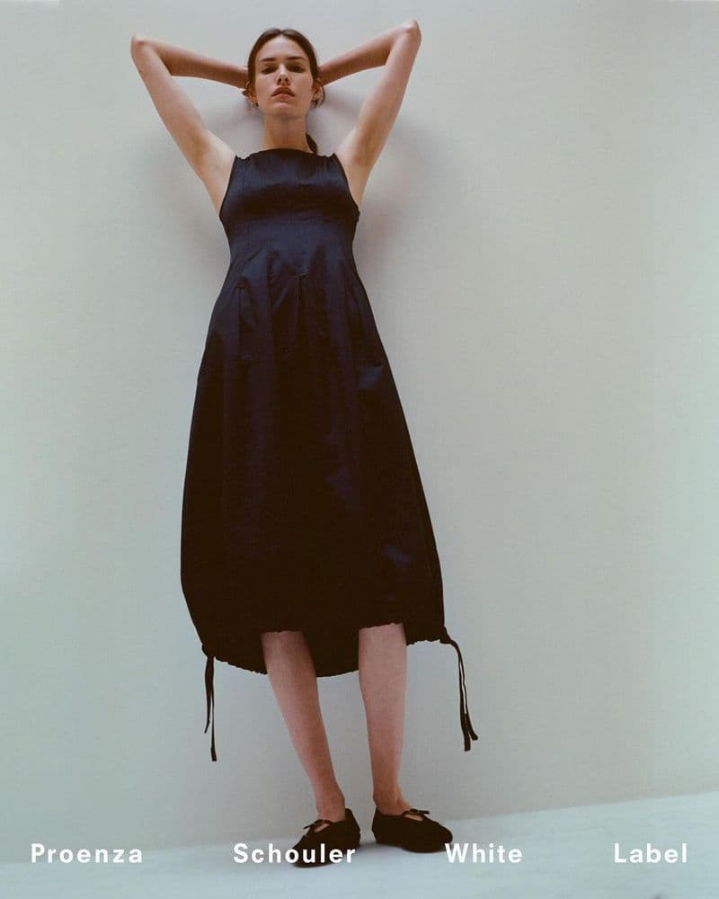Model Vanessa Moody wearing a dark sleeveless midi dress with both hands raised behind her head against a seamless white backdrop