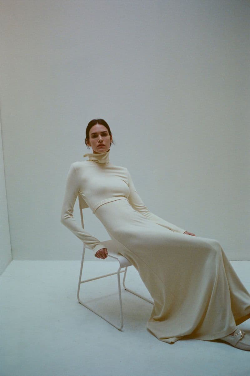 Model wearing a cream long-sleeve maxi dress seated on a white metal chair against a seamless white background