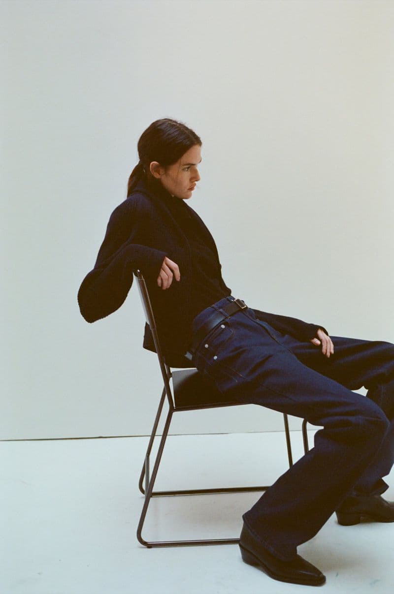 Model Vanessa Moody sitting backward on a metal chair wearing dark blue jeans and a black sweater against a plain white seamless backdrop