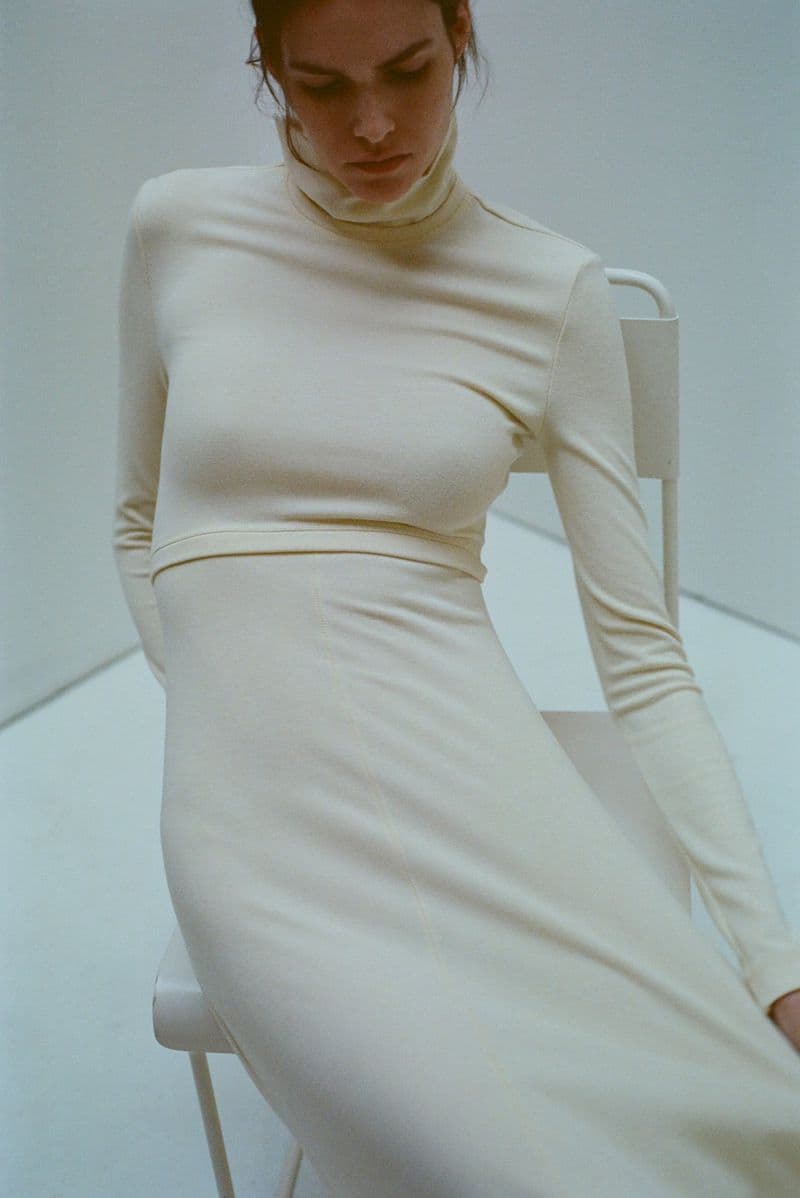 Model Vanessa Moody wearing a cream turtleneck dress seated on a white chair against a plain studio background.