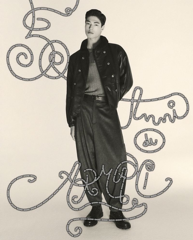 Asian male model in a black leather jacket and baggy pinstripe pants poses in a full shot against a chain-link graphic backdrop.