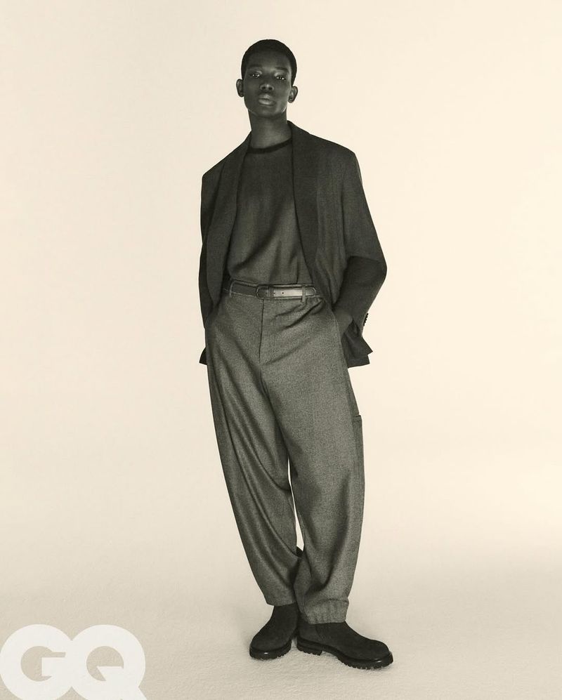 Male fashion model stands in a photo studio wearing a dark blazer, t-shirt, oversized trousers, and dress shoes against a plain backdrop.