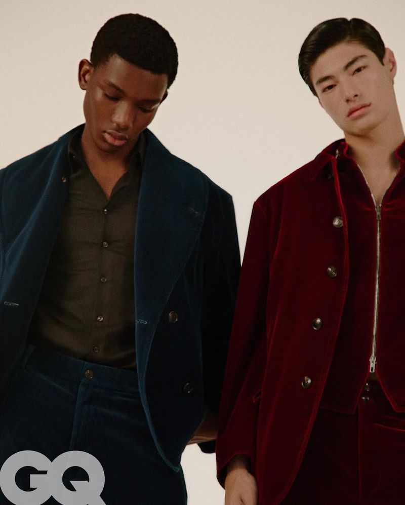 Two adult fashion models in a photo studio pose in velvet suits. The model on the left wears a double-breasted dark blue velvet suit and the model on the right wears a zipper-front burgundy velvet suit.
