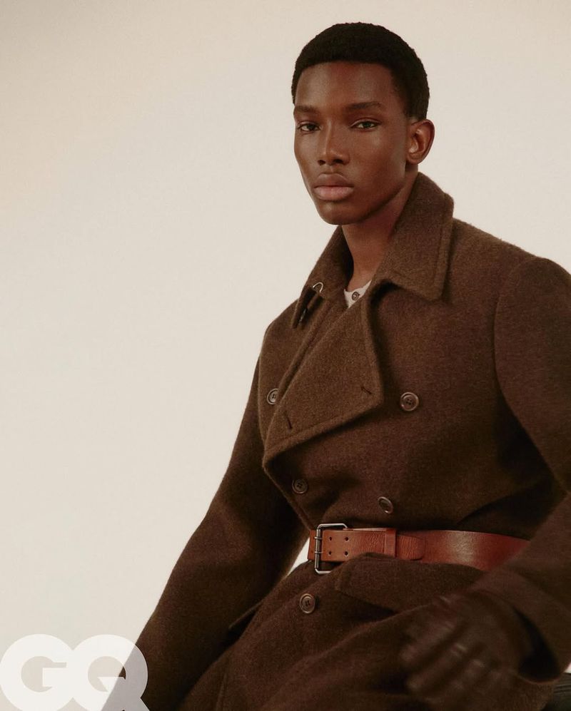 Portrait of a dark-skinned male model in a brown overcoat, wearing a brown belt and gloves, posing indoors against an off-white background.