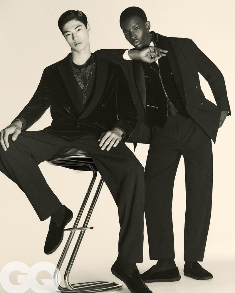Two fashion models in black suits pose indoors; one sits on a chrome stool, while the other leans behind in formal attire.