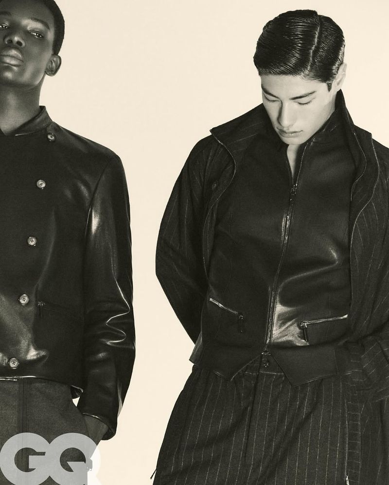 Two men wearing black leather jackets and striped pants pose in a studio shot for a fashion editorial, classic pose and neutral background.
