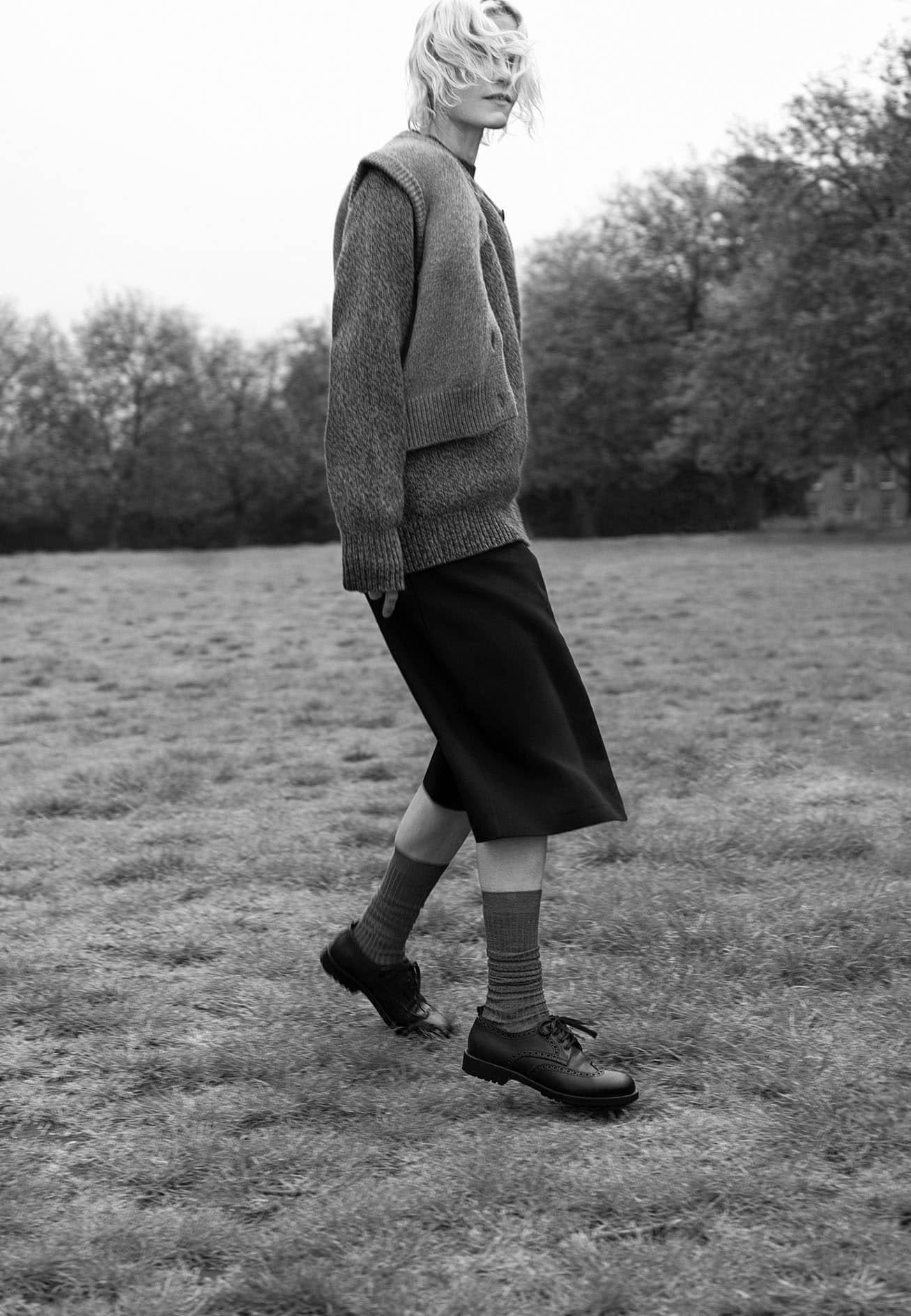 Model walking across a grassy field wearing a textured knit sweater, dark skirt, socks, and lace-up shoes.
