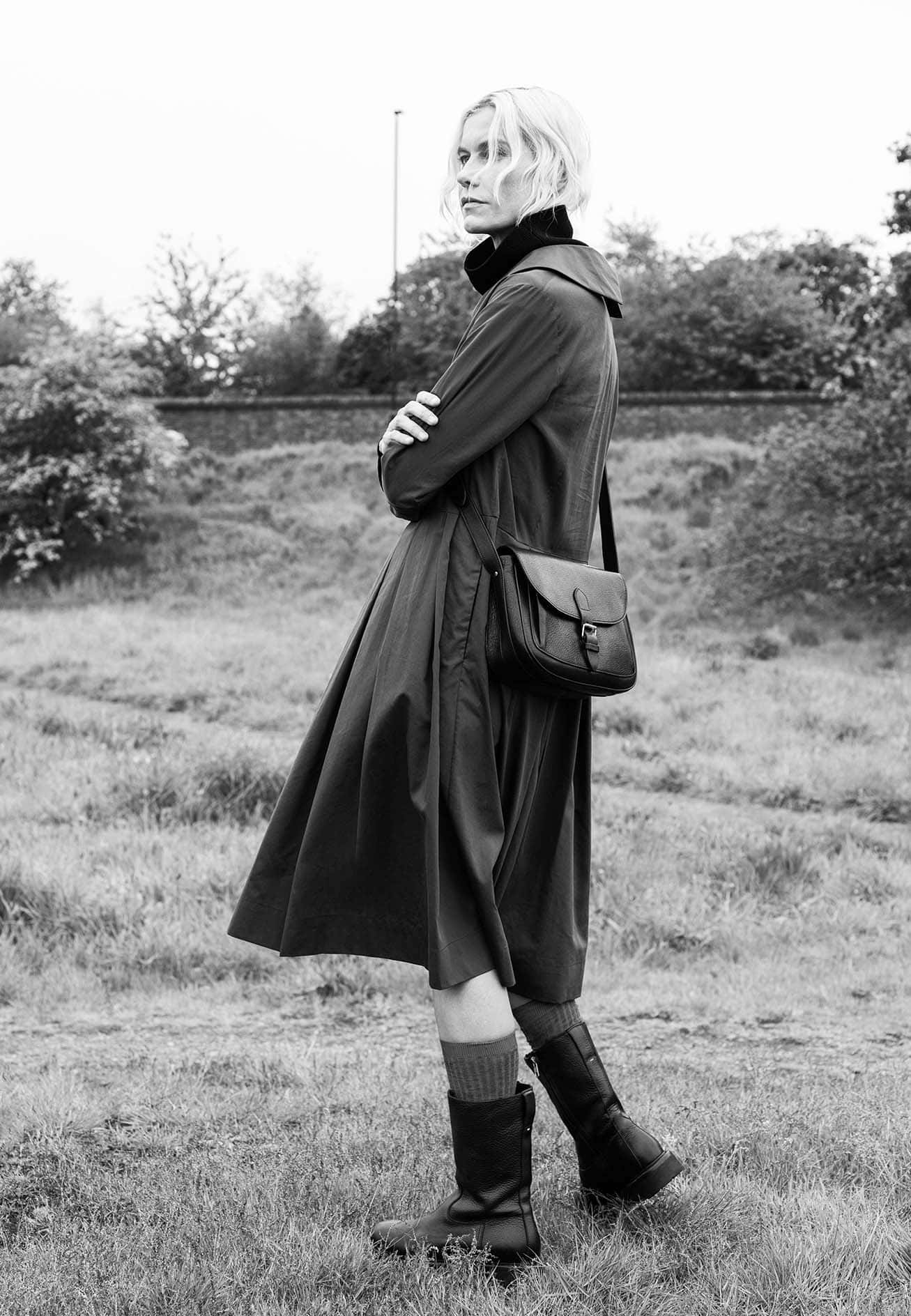 Model standing outdoors in a grassy park wearing a dark belted coat and boots, holding a leather shoulder bag