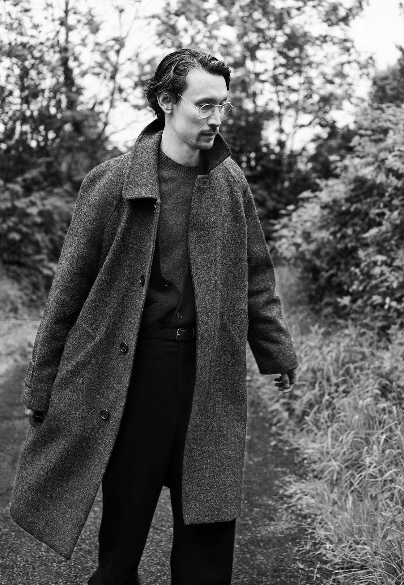 Man wearing a long dark tweed coat and round spectacles standing outdoors on a path bordered by trees and tall grass.
