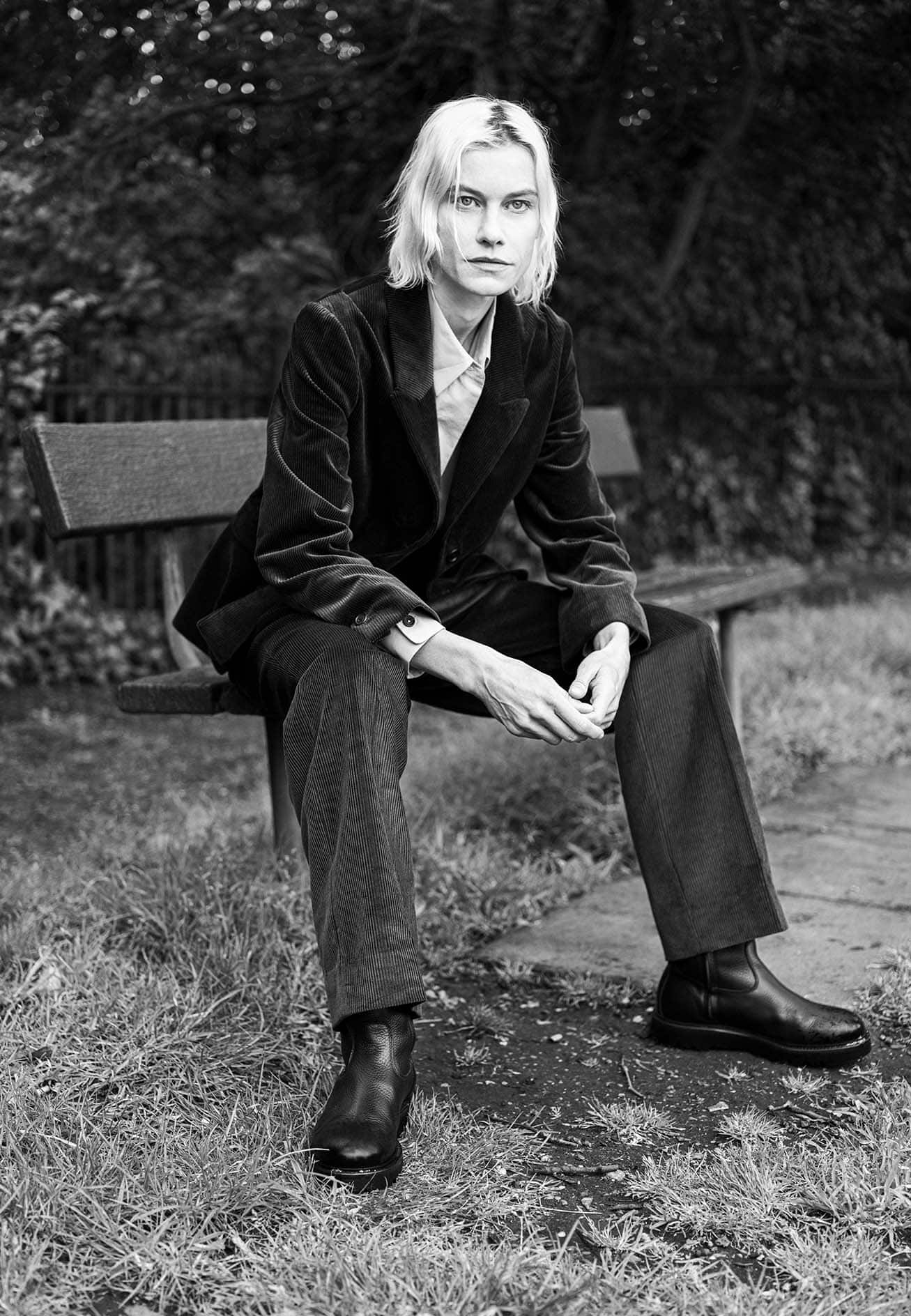 Model wearing a dark velvet blazer and trousers seated on a park bench outdoors