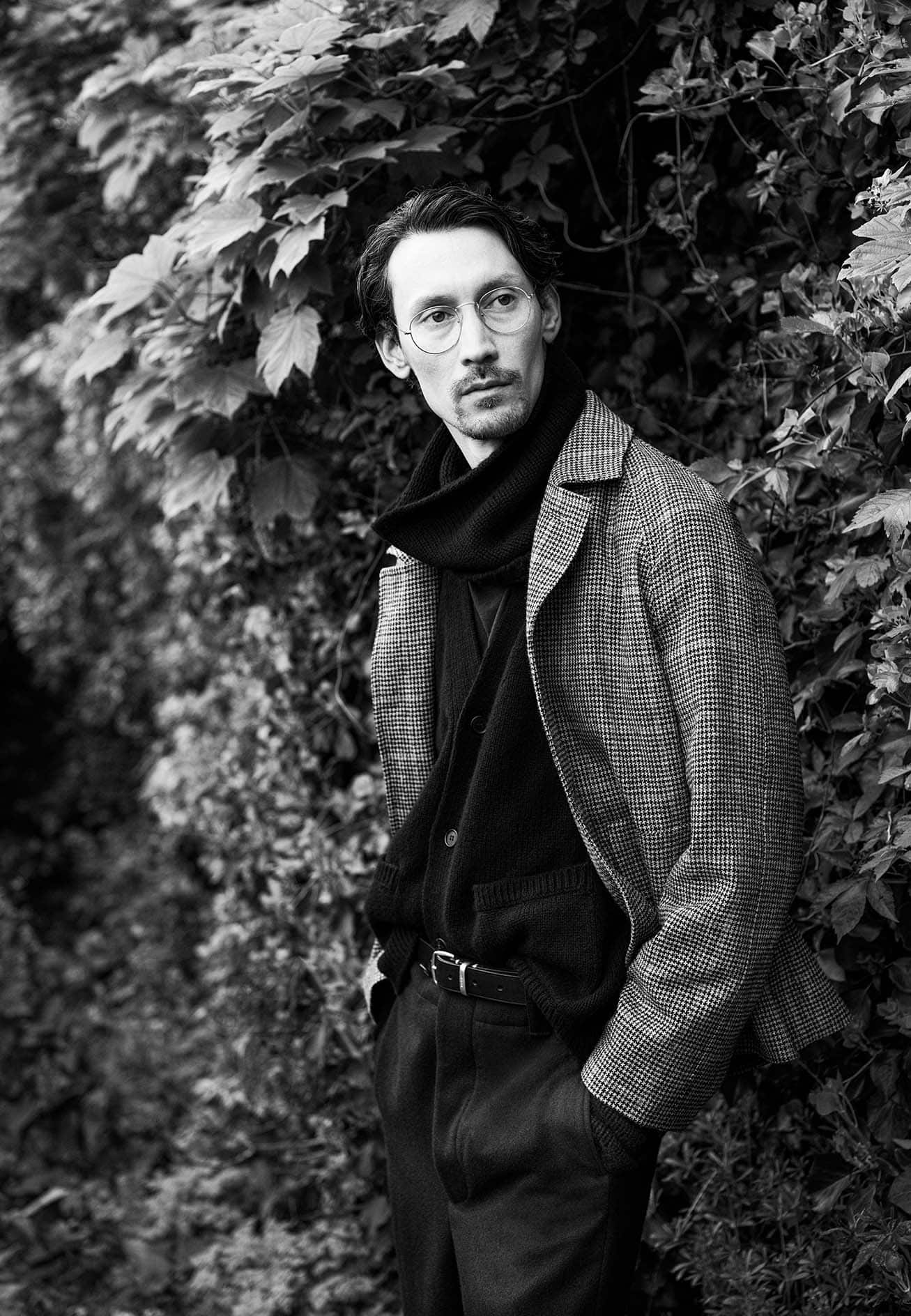 Monochrome portrait of a man in round glasses and a patterned coat standing before dense foliage.