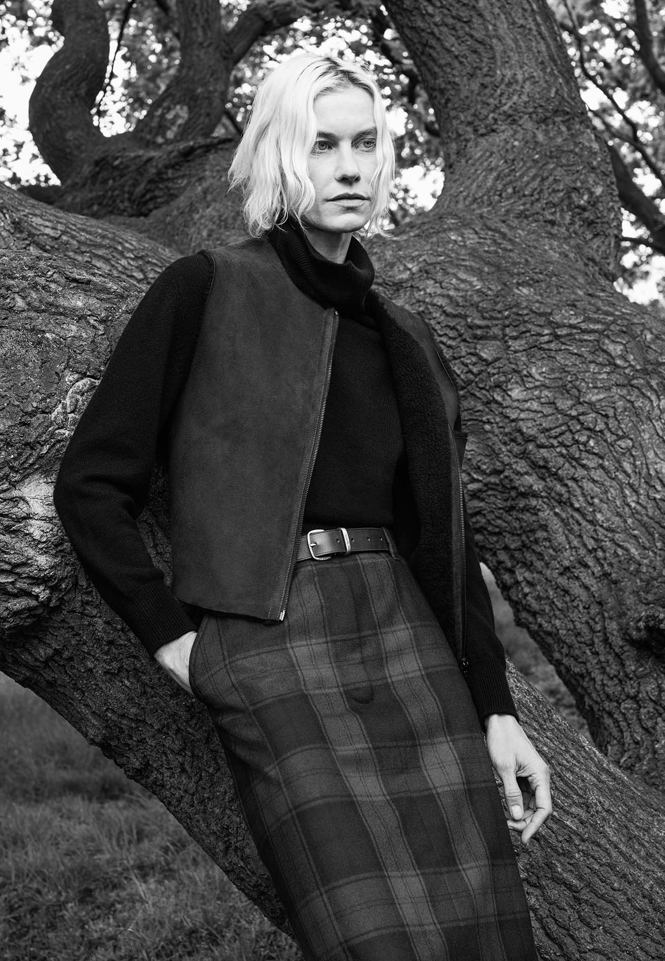 Model with blonde hair leaning against a large tree wearing a dark turtleneck and patterned skirt for Margaret Howell.