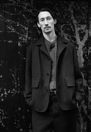 Man wearing a dark jacket and glasses posing against a dark weathered wooden background