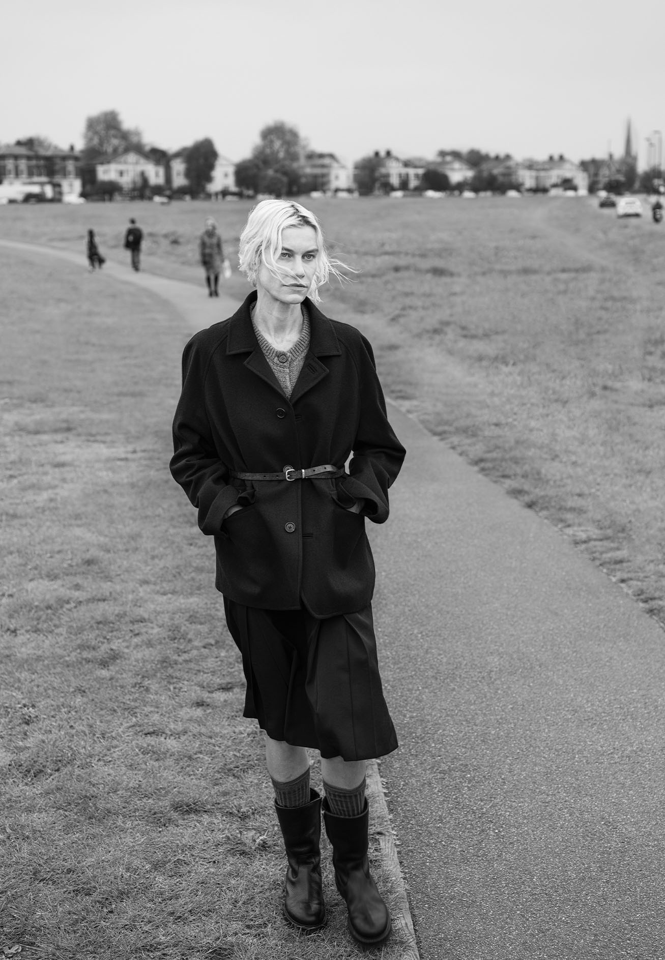 Model walking on an outdoor path wearing a dark jacket, pleated skirt, and boots under an overcast sky.