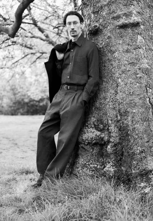 Monochrome portrait of a man leaning against a large tree trunk in an outdoor park setting, wearing a button-down shirt and trousers.