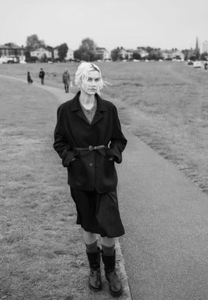 Model walking on an outdoor path wearing a dark jacket, pleated skirt, and boots under an overcast sky.