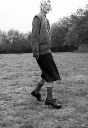 Model walking across a grassy field wearing a textured knit sweater, dark skirt, socks, and lace-up shoes.