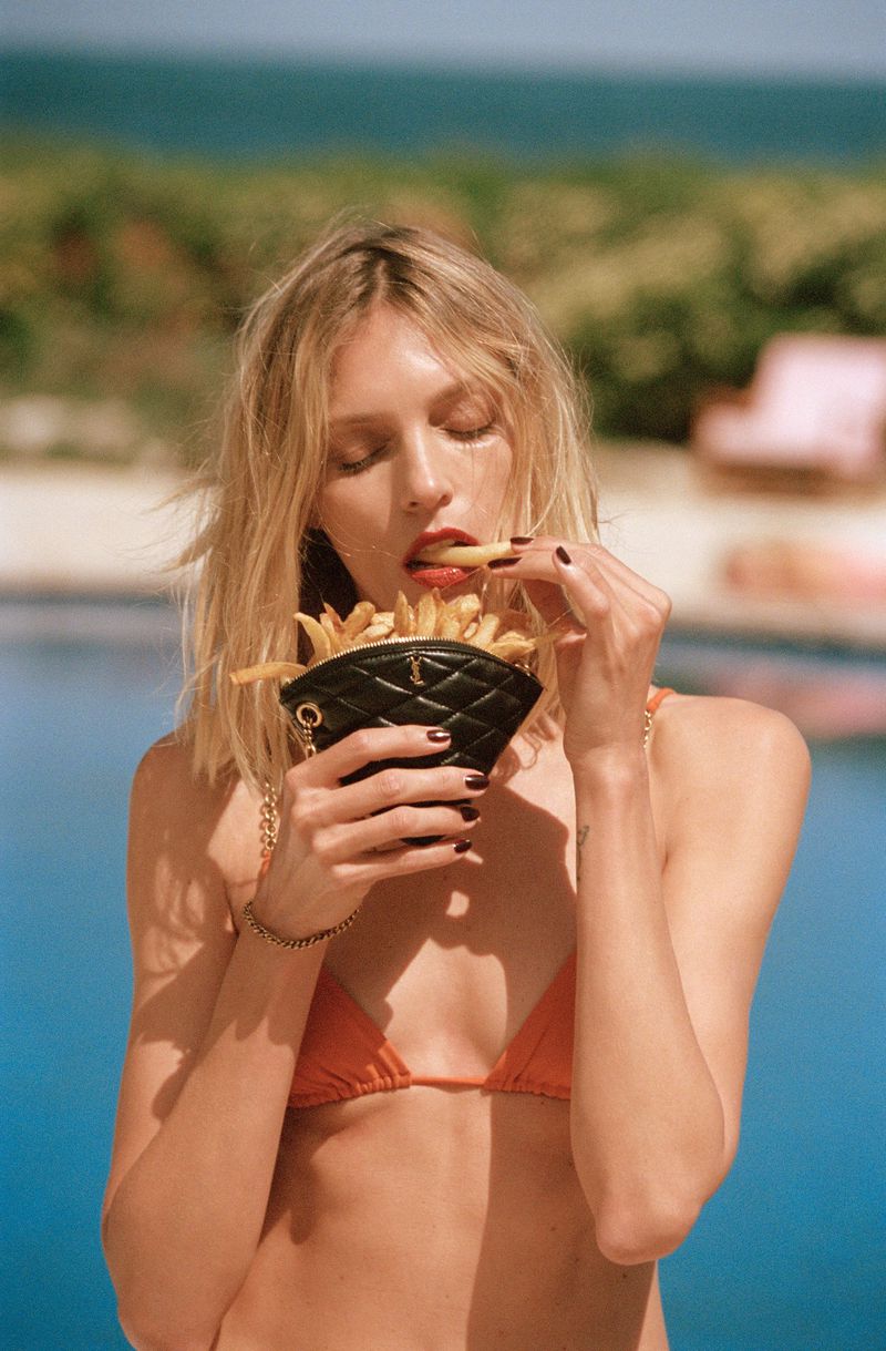 Anja Rubik in an orange bikini holding a quilted black bag eating fries by a pool for Saint Laurent Tan Lines Summer 2024 campaign