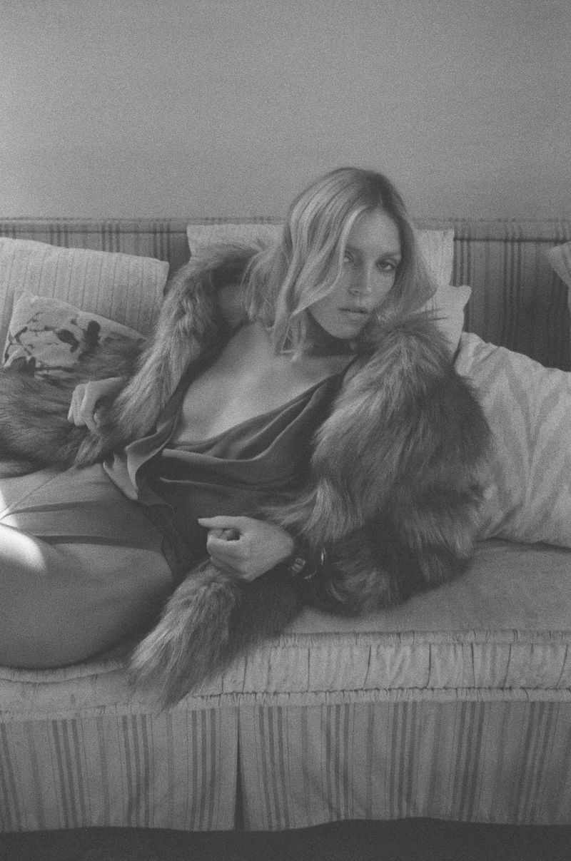 Anja Rubik reclining on a striped sofa wearing a dark slip dress and fur stole for Saint Laurent Tan Lines campaign.