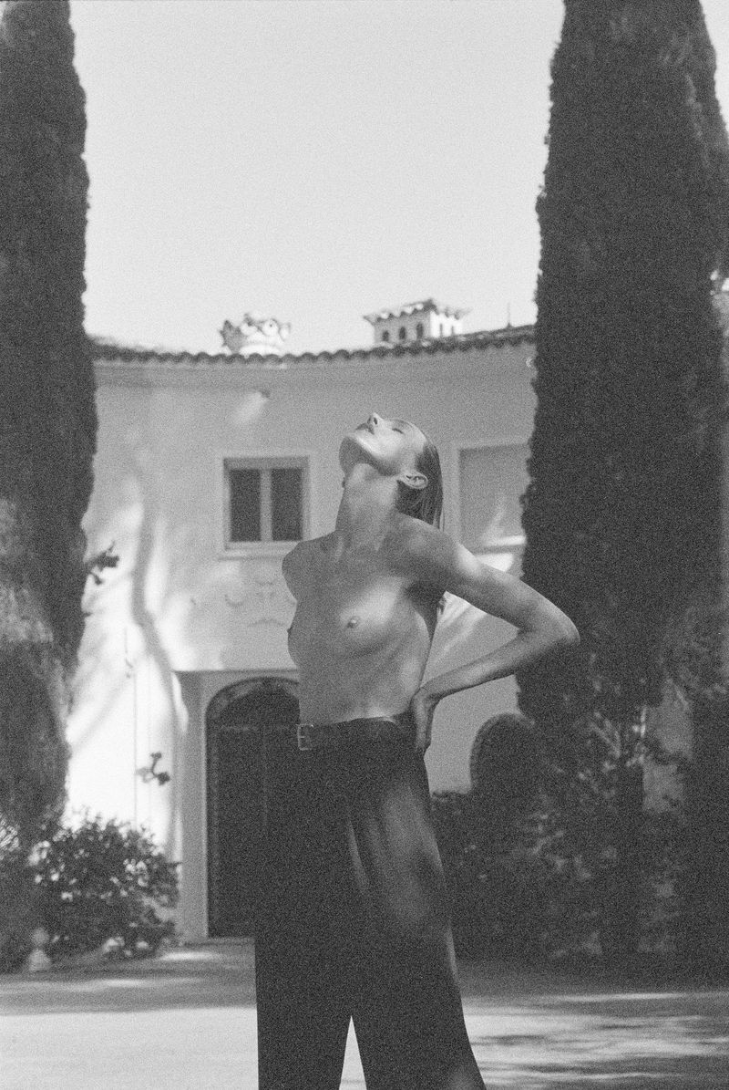Model Anja Rubik posing outdoors in dark trousers with tall cypress trees framing a classic building in background