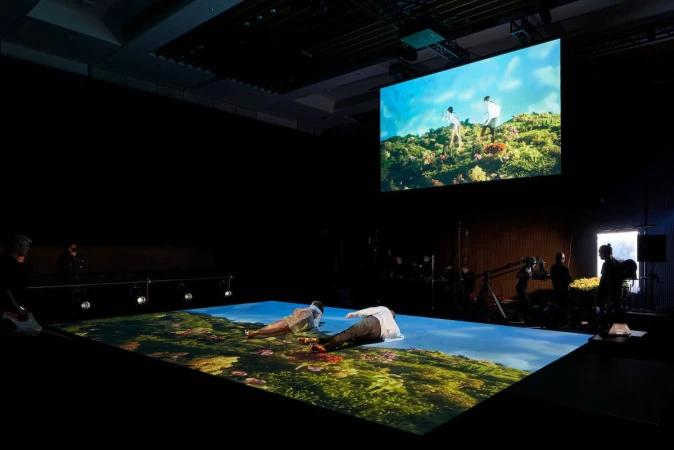 Behind the scenes shot of two performers lying on a floor projection screen inside a dark stage studio with camera crew.