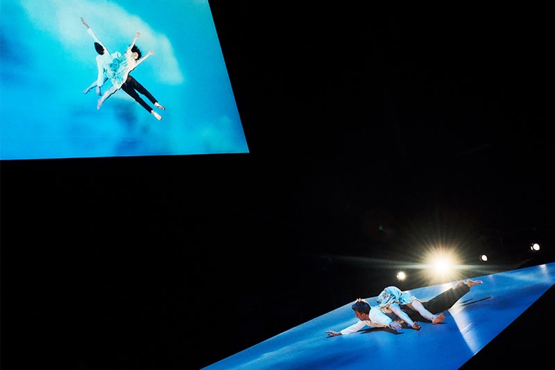 Two figures performing choreography on a tilted blue stage beneath a suspended screen showing a mirrored aerial reflection.