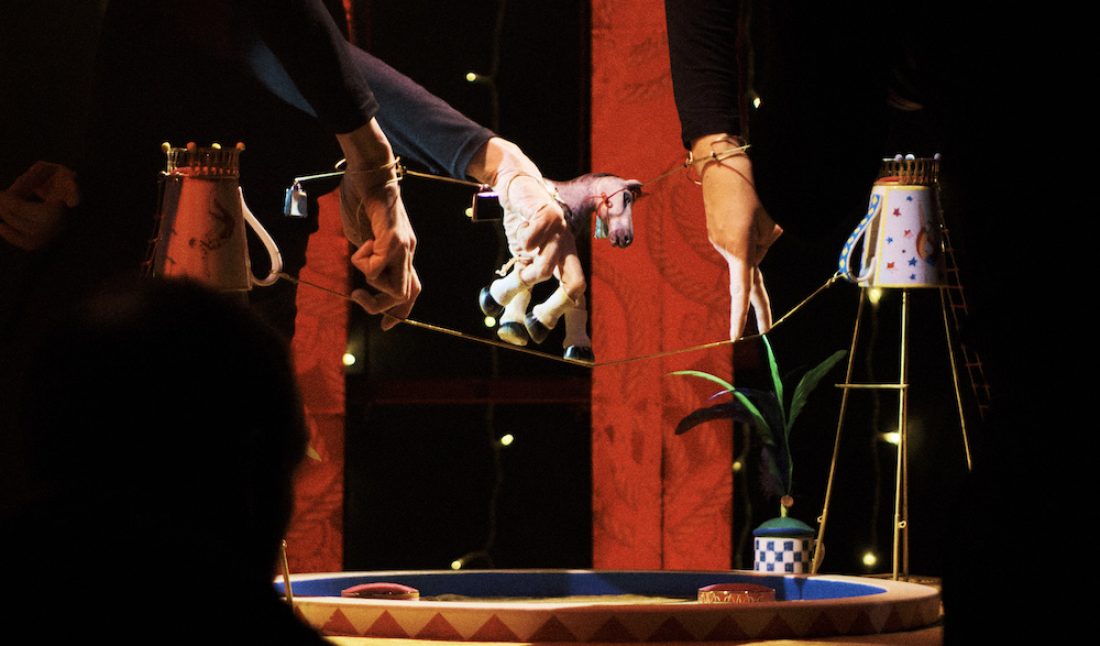 Puppeteers' hands manipulate a miniature toy horse walking tightrope above a circular circus ring stage set