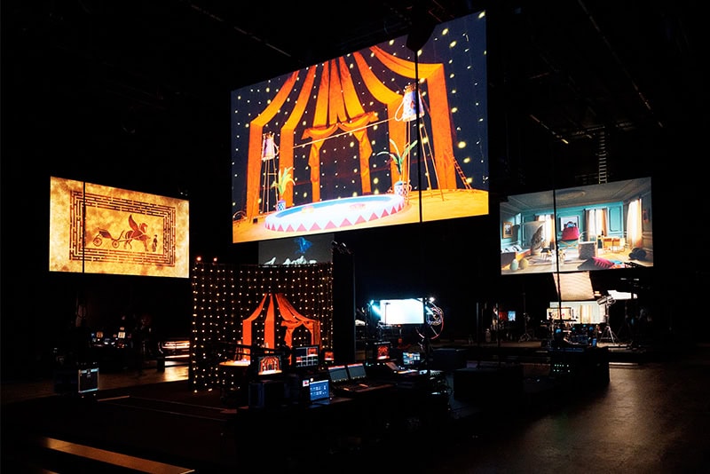 Large dark soundstage illuminated by multiple glowing screens displaying circus and theatrical stage sets for Hermes ad campaign