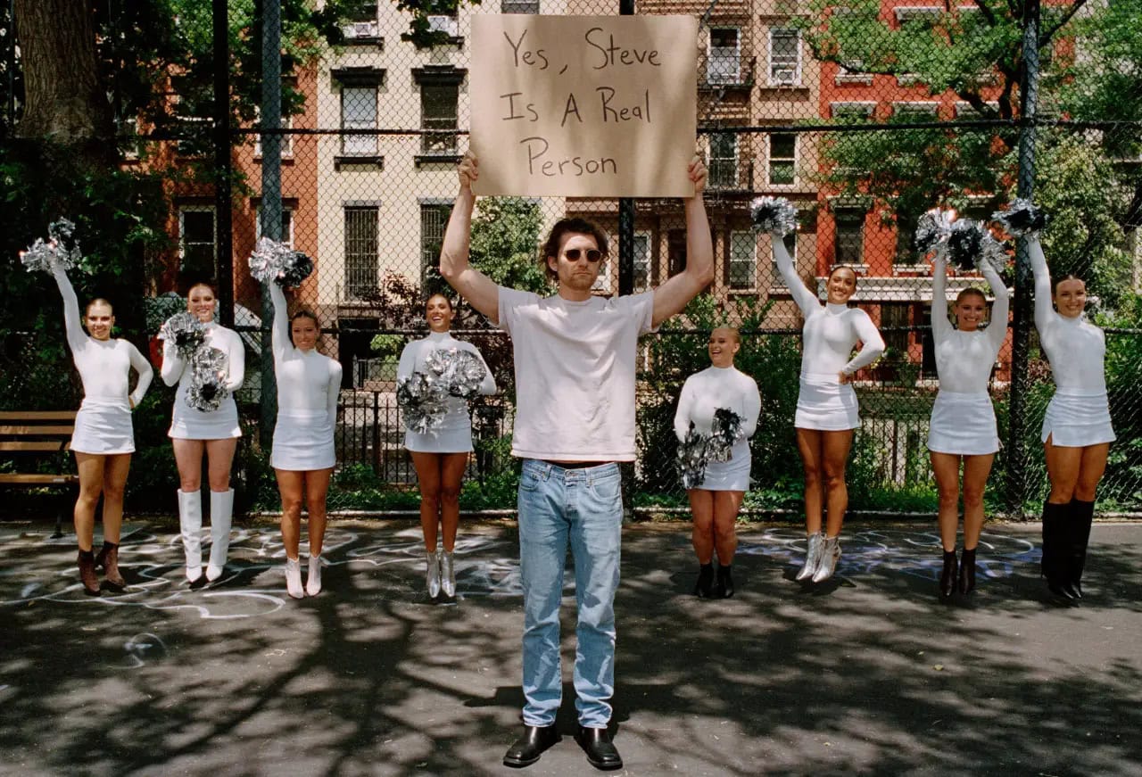 Man holding a cardboard sign reading Yes, Steve Is A Real Person framed by cheerleaders holding pom poms outdoors