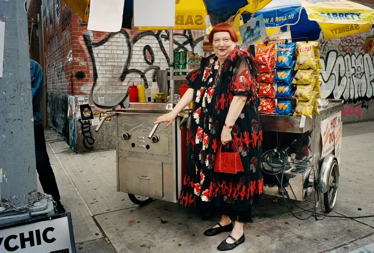 An elderly woman with bright red hair standing next to a New York City hot dog cart holding a red handbag