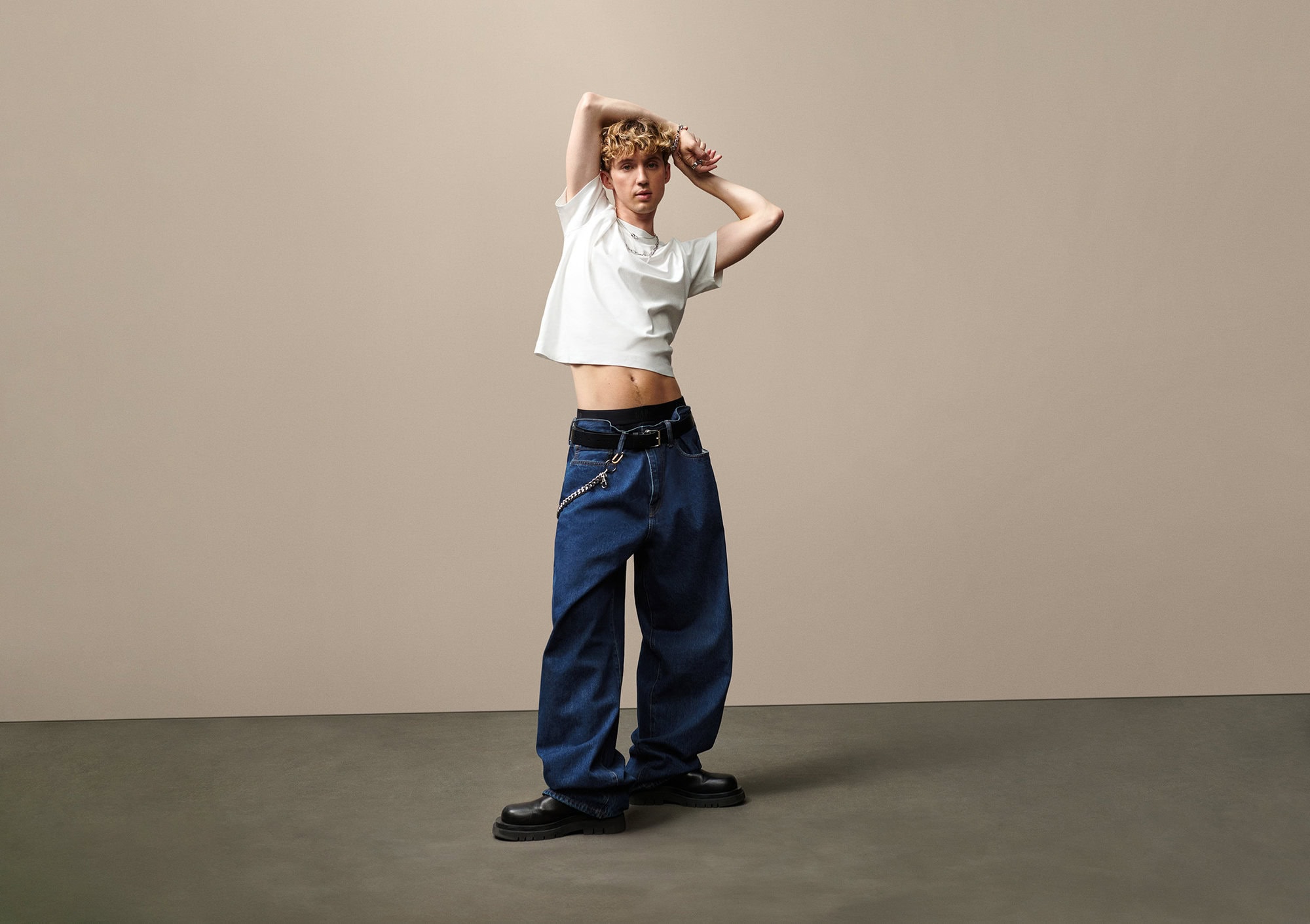Troye Sivan posing in a white t-shirt and wide-leg denim jeans against a seamless neutral studio backdrop.