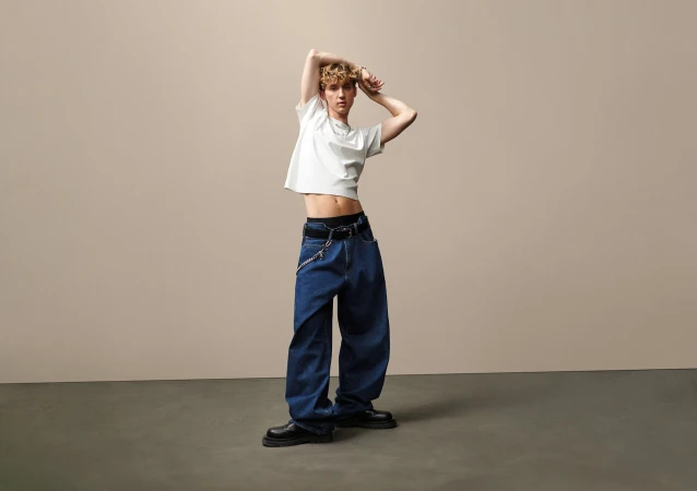 Troye Sivan posing in a white t-shirt and wide-leg denim jeans against a seamless neutral studio backdrop.