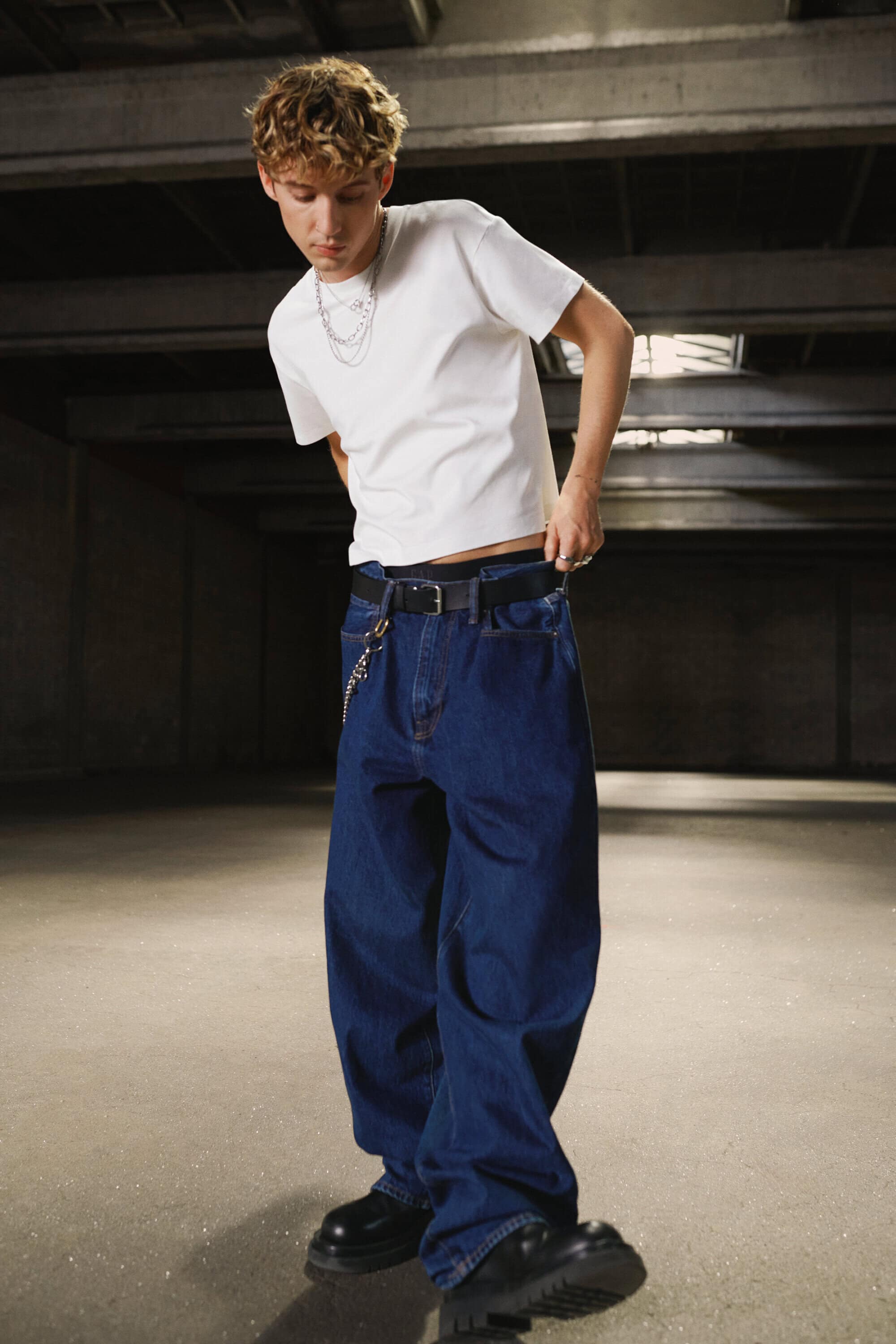Troye Sivan wearing a white t-shirt and loose dark denim jeans in an industrial warehouse setting for Gap