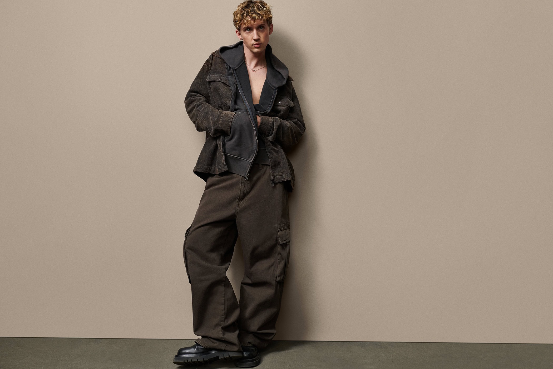 Troye Sivan leaning against a plain wall wearing a dark brown corduroy jacket and matching cargo pants for Gap Get Loose campaign