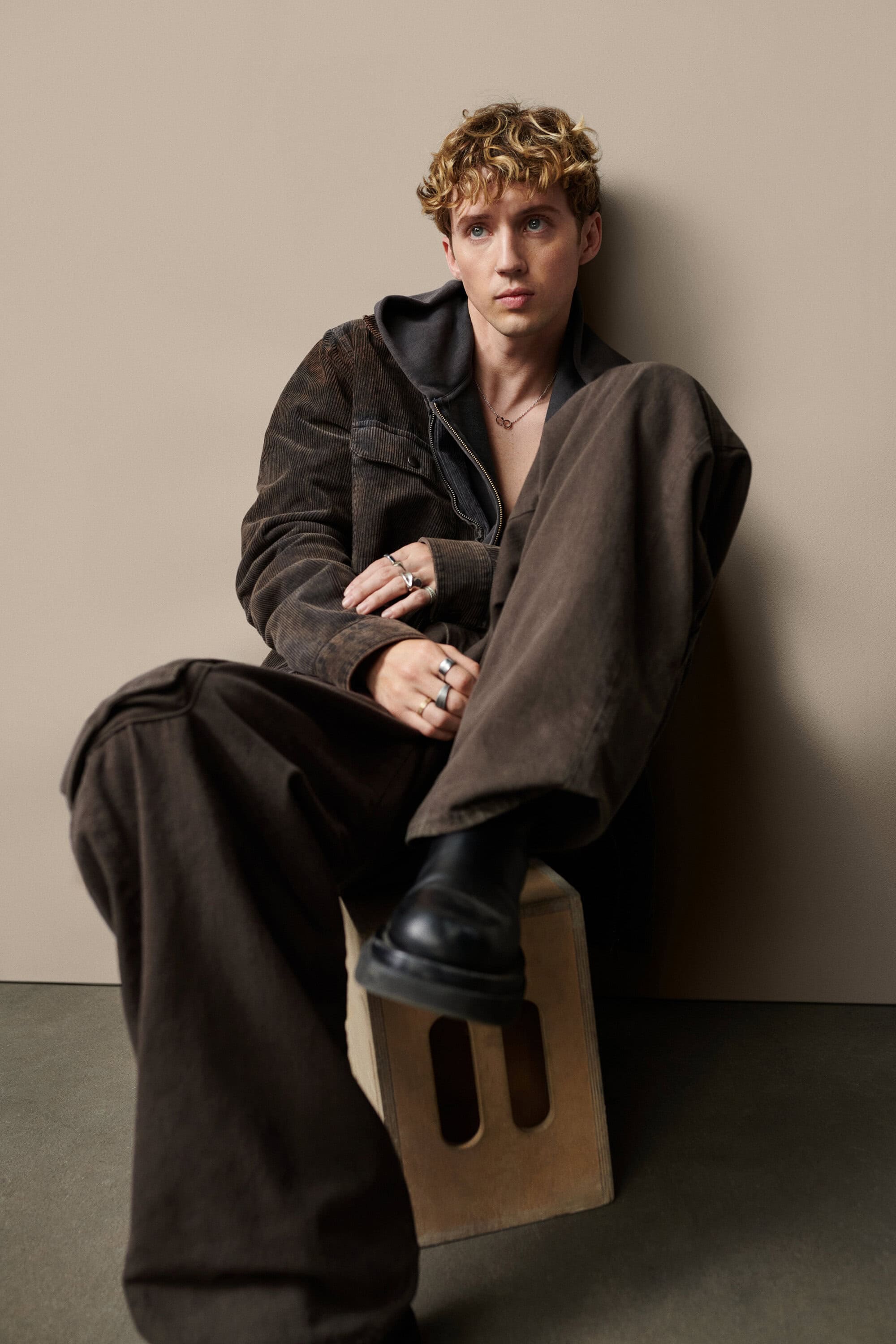 Troye Sivan seated on a wooden crate wearing a brown jacket and wide-legged trousers against a plain studio background.
