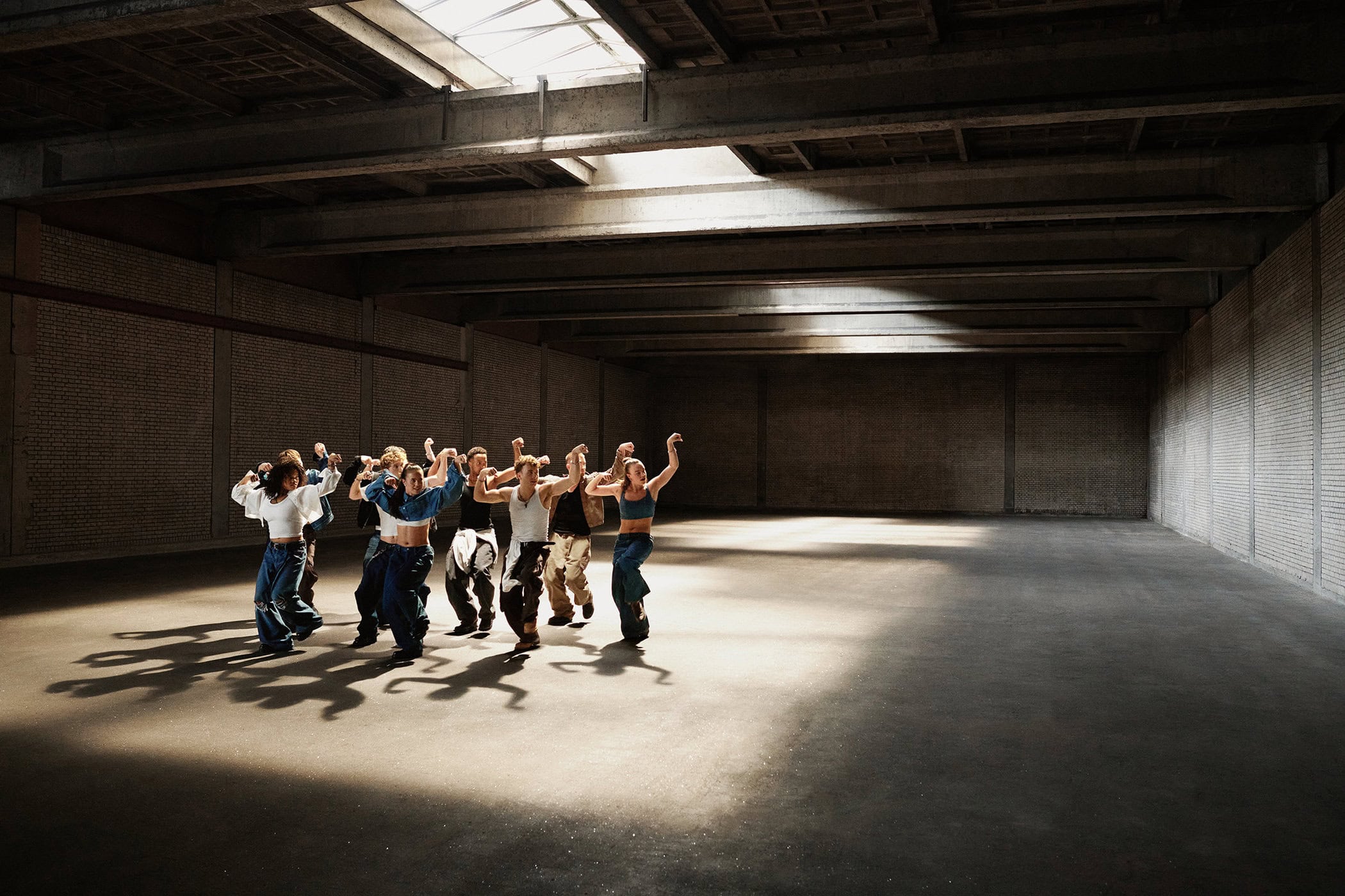 A group of dancers performing in a large empty warehouse under natural sunlight streaming from a ceiling skylight.