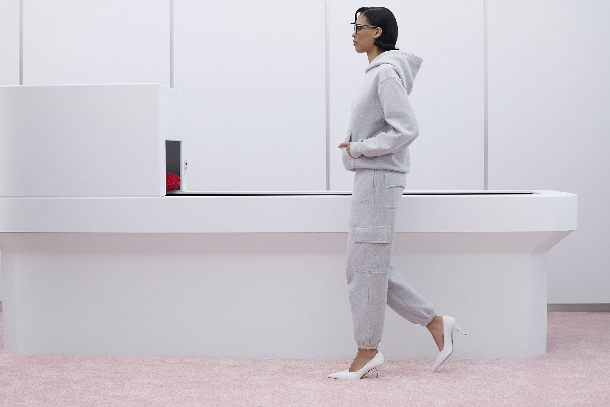 Model Nara Aziza Smith walking in a light grey hoodie and sweatpants with white heels against a minimalist white set.