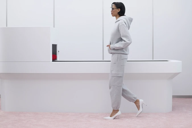 Model Nara Aziza Smith walking in a light grey hoodie and sweatpants with white heels against a minimalist white set.