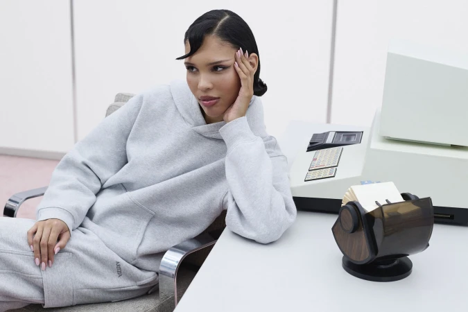 Model Nara Aziza Smith wearing a grey hoodie in an office setting for the Aritzia Sweatfleece campaign.