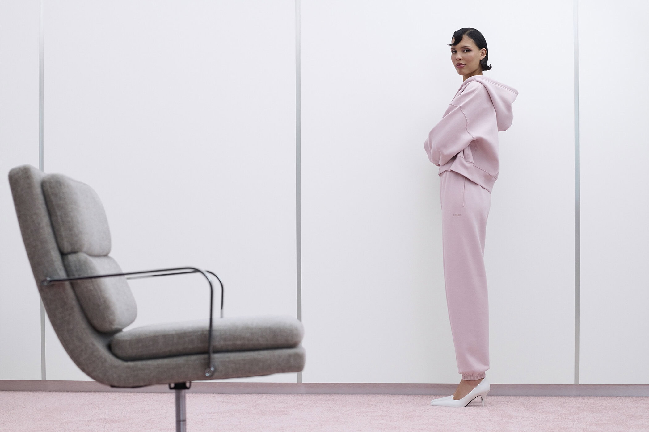 Model Nara Aziza Smith standing in pink sweatsuit beside a grey armchair against a pale paneled wall for Aritzia.