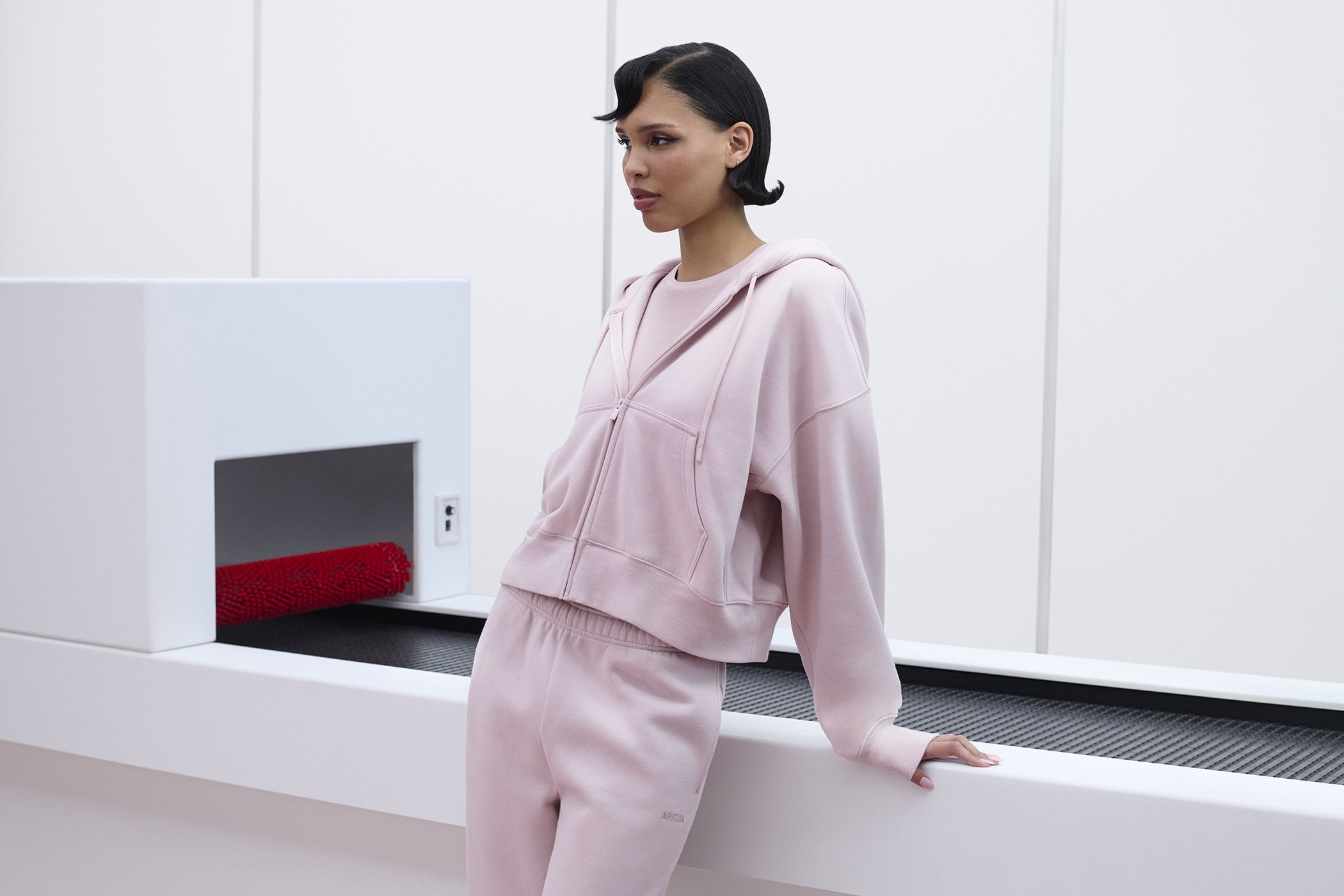 Model Nara Aziza Smith wearing a pink zip hoodie and matching sweatpants leaning against a white conveyor structure for Aritzia.