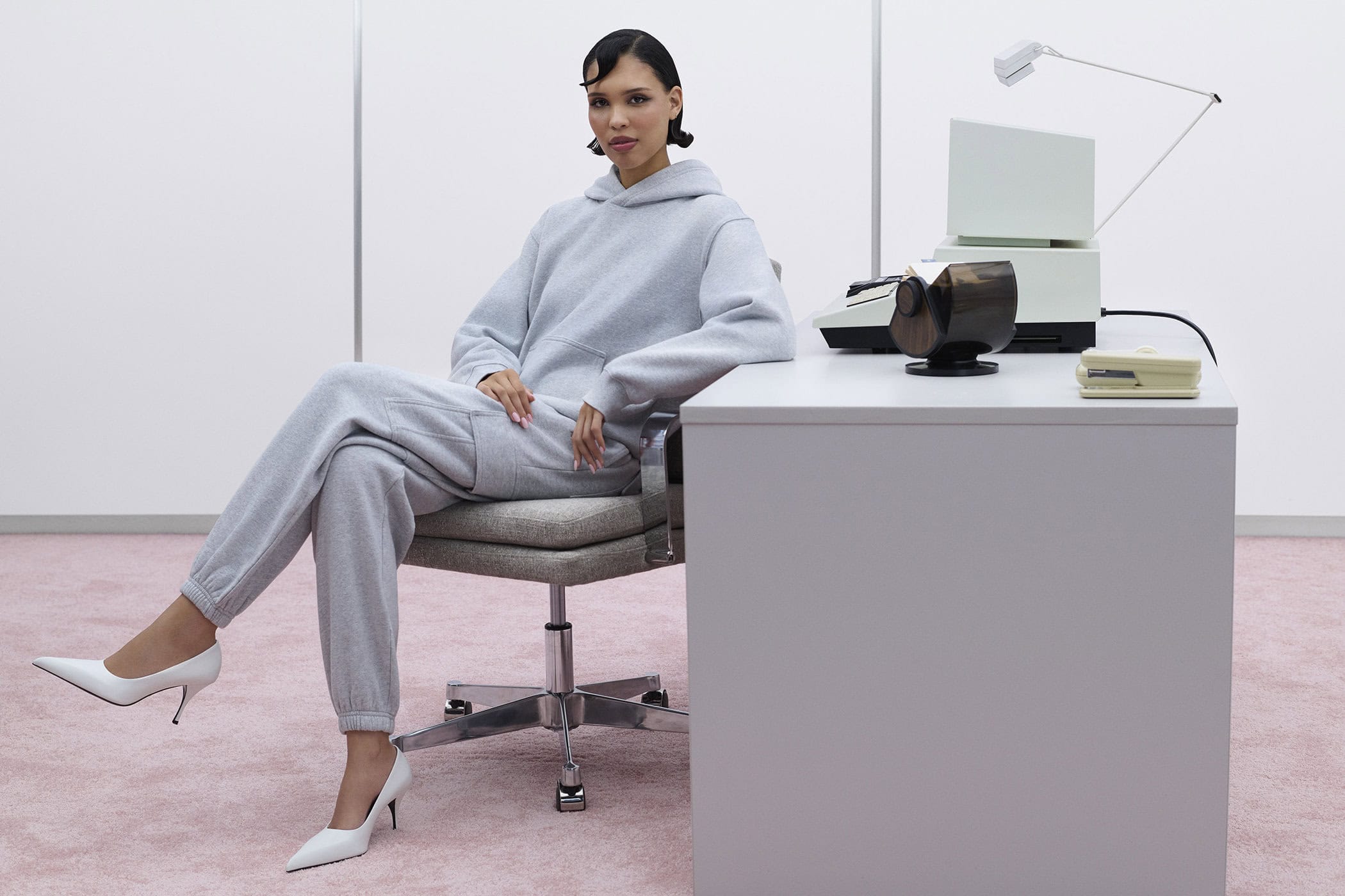 Model Nara Aziza Smith in a grey sweatfleece hoodie and matching sweatpants seated at a modern office desk for Aritzia.