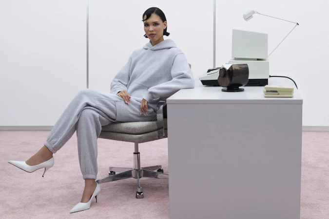 Model Nara Aziza Smith in a grey sweatfleece hoodie and matching sweatpants seated at a modern office desk for Aritzia.
